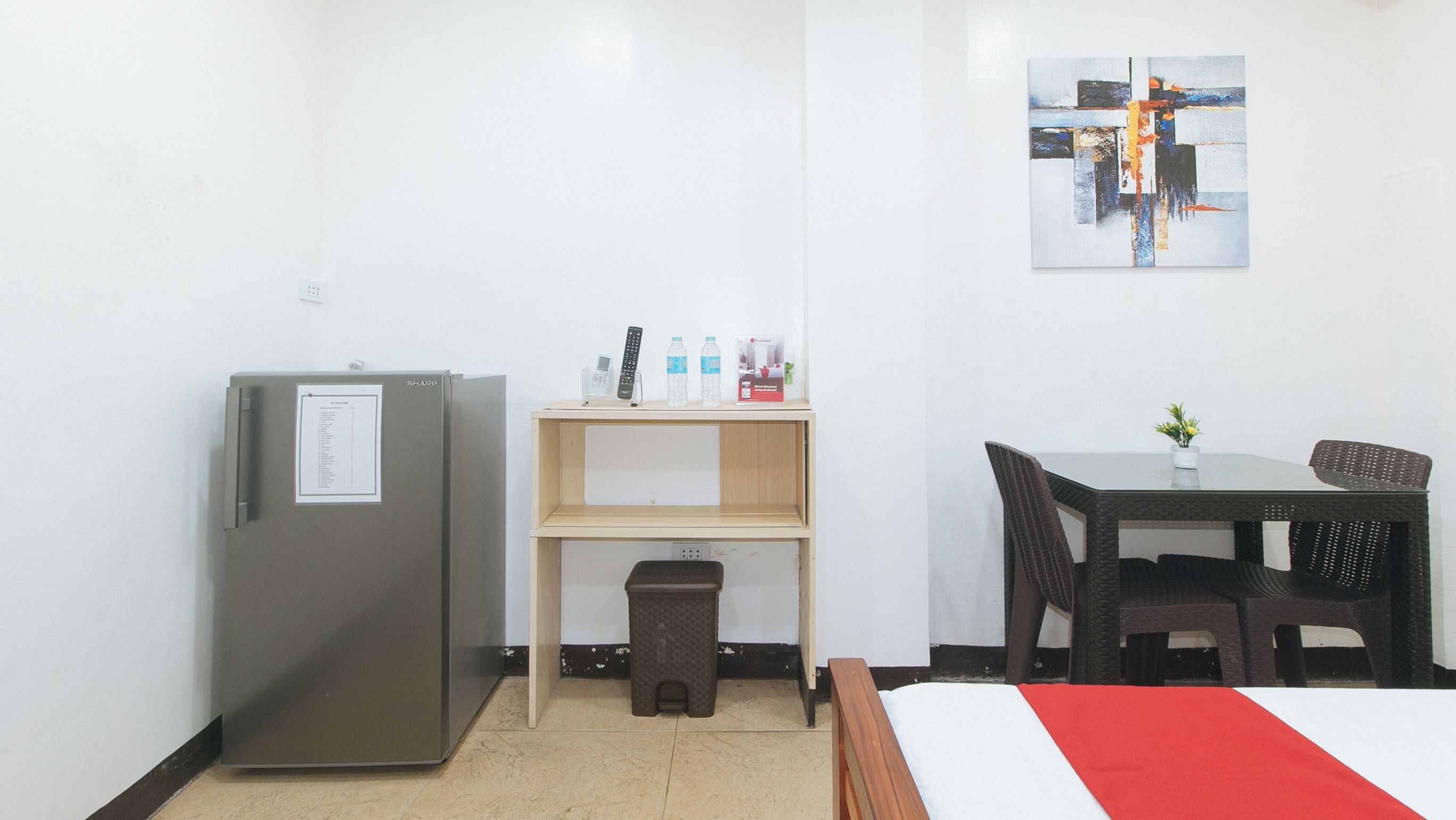 A hotel room with a bed, a desk with a chair, a refrigerator, a trash bin, and a shelf with a remote control, water bottles, and a box.