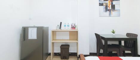 A hotel room with a bed, a desk with a chair, a refrigerator, a trash bin, and a shelf with a remote control, water bottles, and a box.