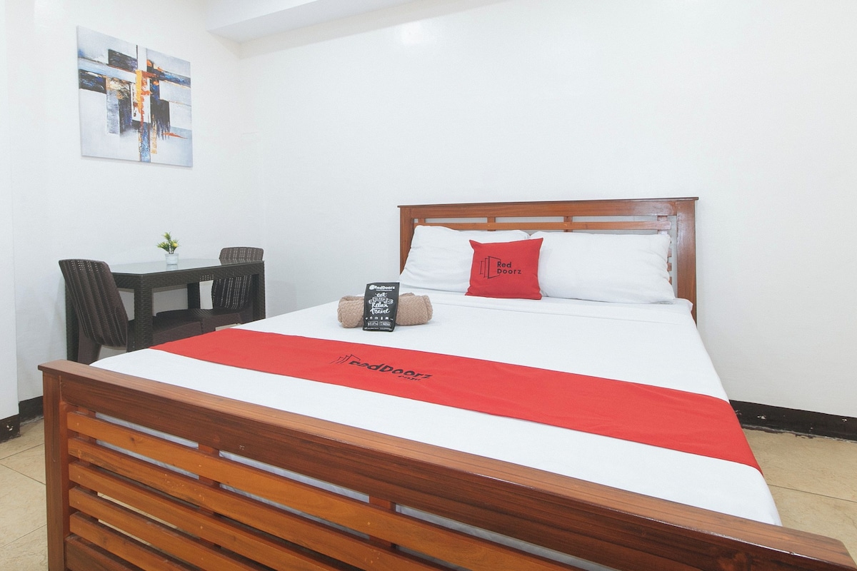 Une chambre d’hôtel avec un lit en bois, du linge de lit blanc, un plaid rouge, un coussin Red Doorz, une petite table avec une plante et un tableau au mur.