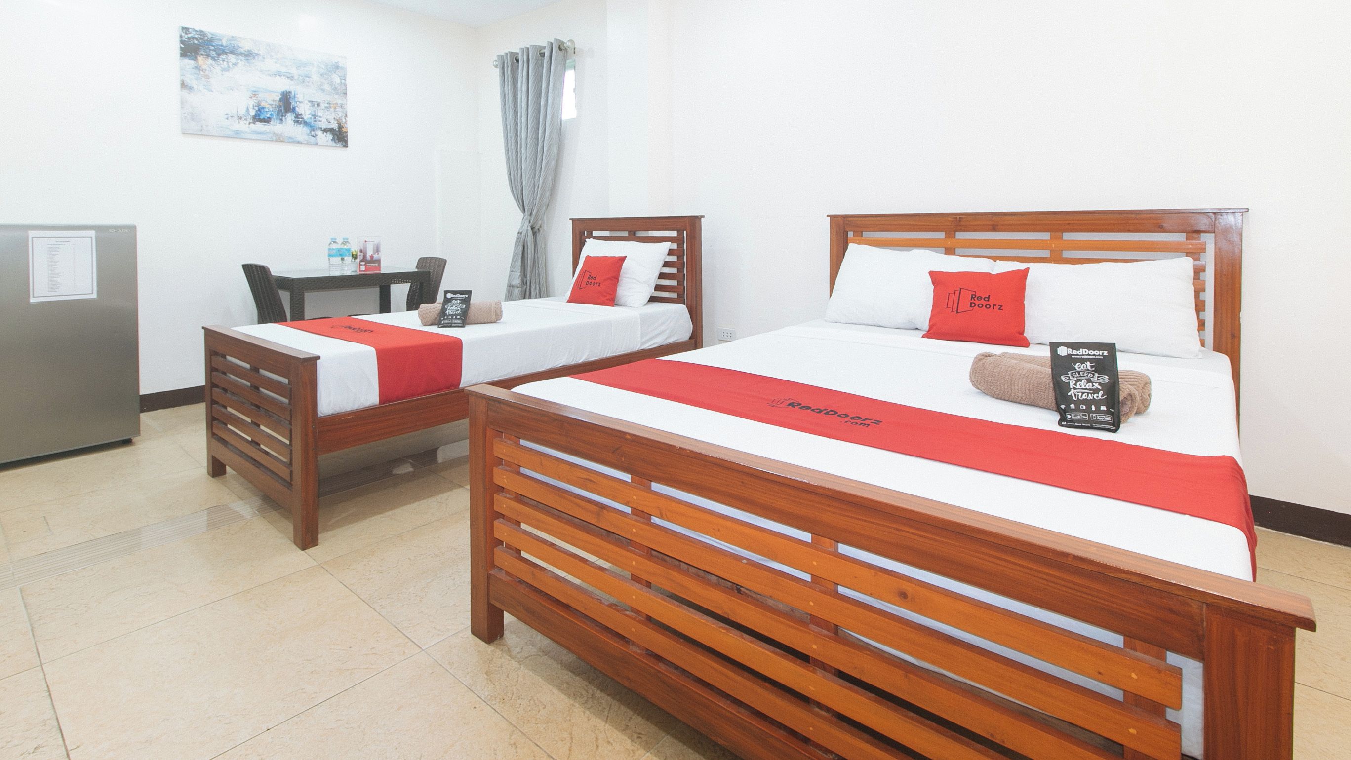 Una habitación de hotel con dos camas de madera, cada una con colcha y almohadas rojas, un escritorio con silla, una mesita pequeña y un refrigerador.
