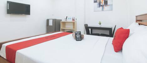 A hotel room with a bed, a desk, a chair, a television, and a RedDoorz sign on the bed.