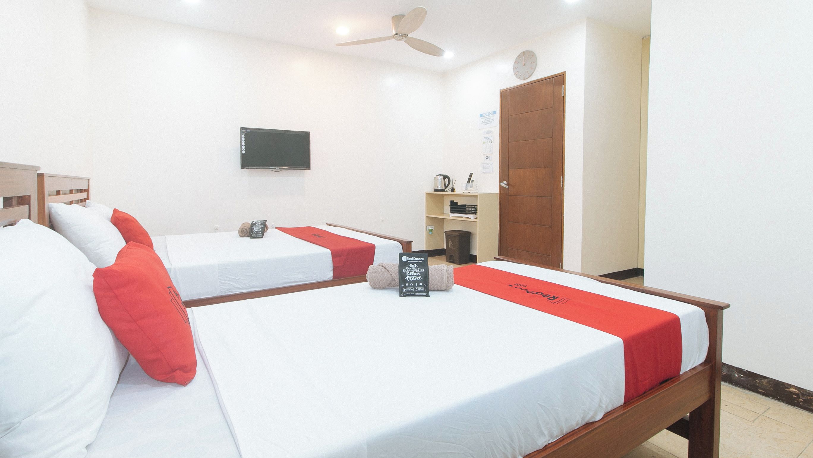 Habitación de hotel con dos camas individuales, un televisor montado en la pared, una puerta de madera y un estante con objetos.