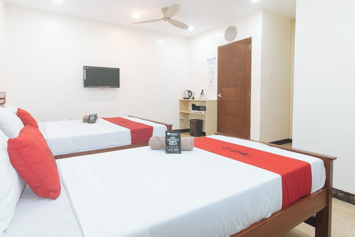 Habitación de hotel con dos camas individuales, un televisor montado en la pared, una puerta de madera y un estante con objetos.