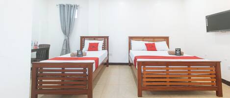 Une chambre d’hôtel avec deux lits en bois, chacun recouvert d’une couverture rouge et agrémenté d’oreillers, une petite table avec une chaise, une fenêtre avec des rideaux et un téléviseur à écran plat fixé au mur.
