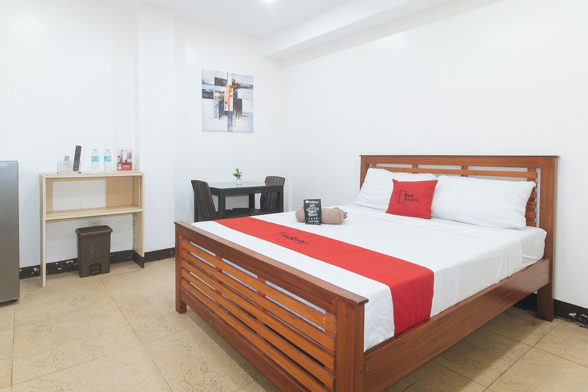 A hotel room with a bed, a wooden headboard, a red pillow, a small table with a plant, a shelf with items, and a refrigerator.