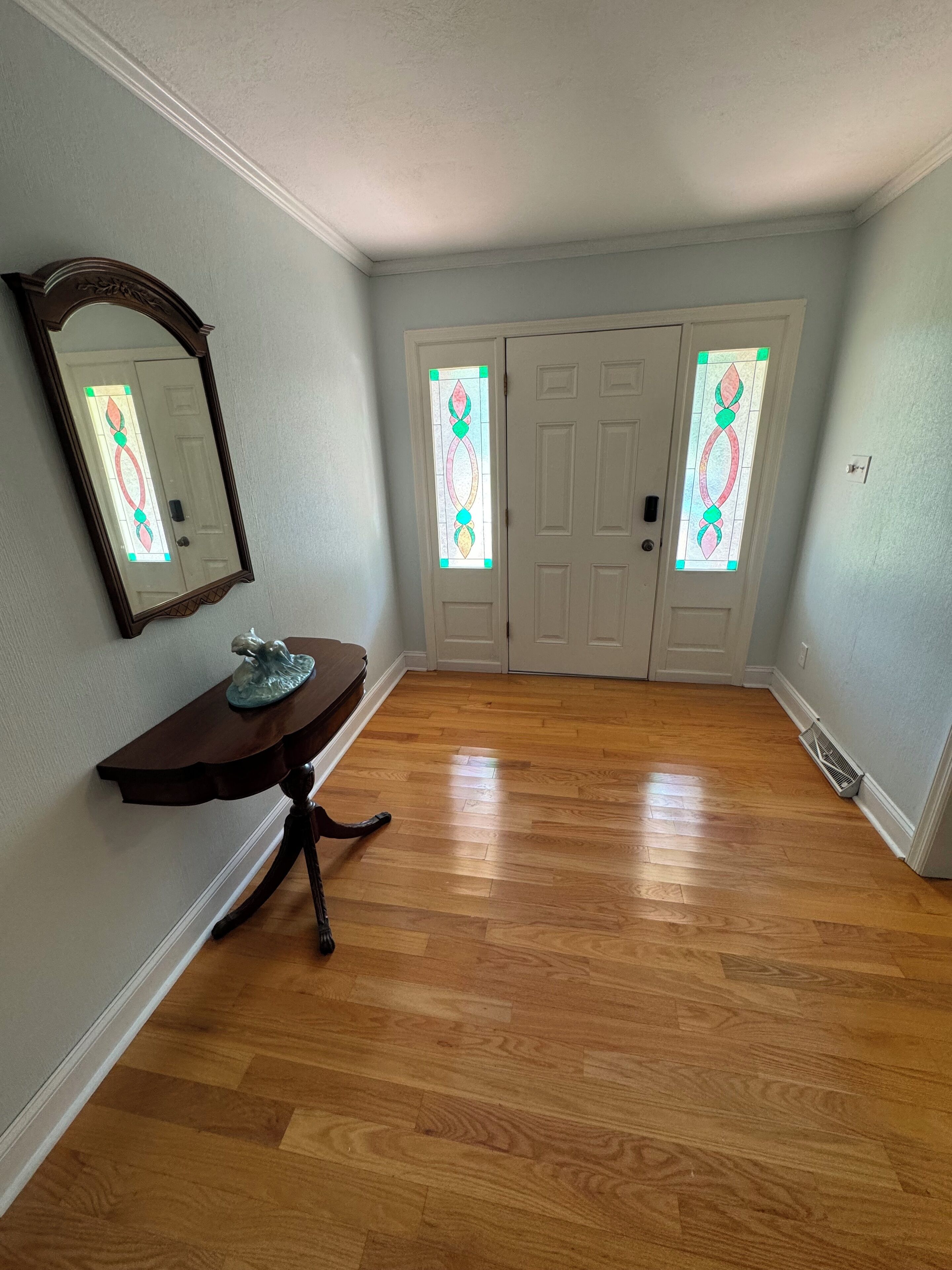 A hallway with wooden flooring, a dark wooden console table with a decorative item, and a mirror on the left wall. There are two white doors with stained glass windows.