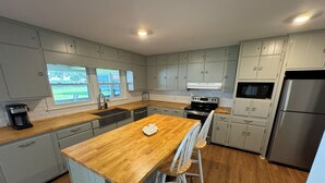 Une cuisine moderne avec des meubles de couleur bleu clair, un îlot central en bois doté de tabourets, des appareils électroménagers en acier inoxydable et une vue sur l’extérieur grâce à de grandes fenêtres.