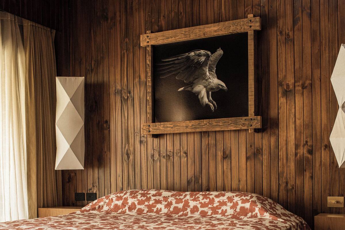 A bedroom with a wooden wall, a bed with a patterned bedspread, a black leather bench, and a wall-mounted artwork of an eagle.