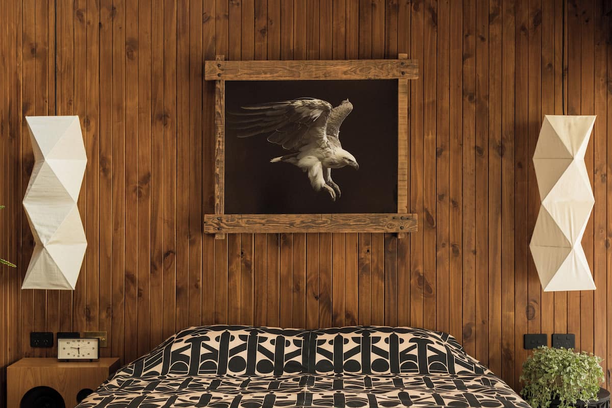 A room with wooden panel walls, a framed picture of an eagle, a bed with patterned bedding, a clock, and a small plant.