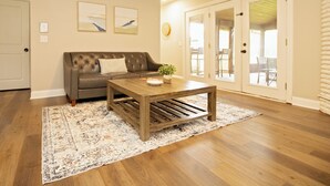 A living room with a brown leather sofa, a wooden coffee table, and a patterned rug. There are two framed pictures on the wall and a door leading to an outdoor area.