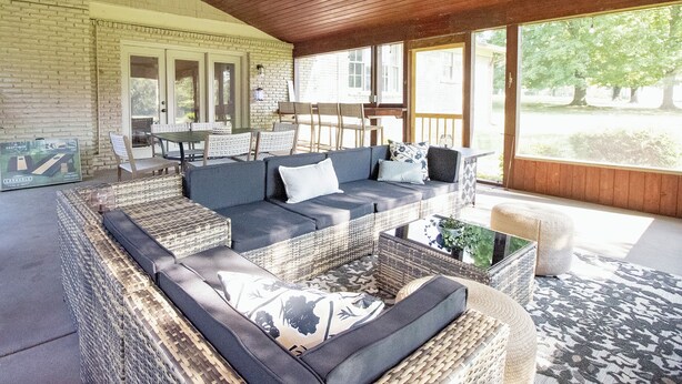 A covered outdoor seating area with wicker furniture, a ceiling fan, and large windows offering a view of the outdoors.