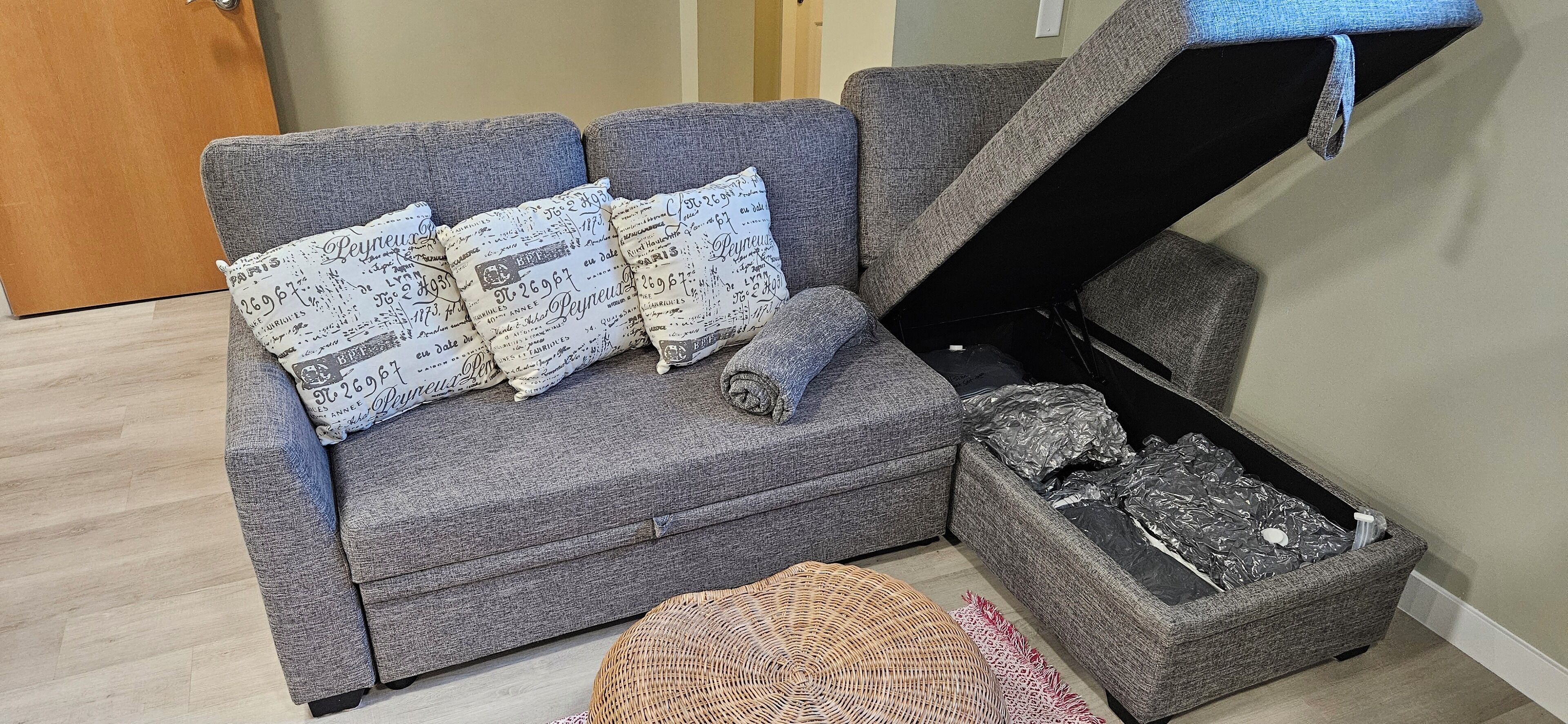 Un sofá gris con almohadones estampados, un pequeño otomaní y un baúl con interior negro.