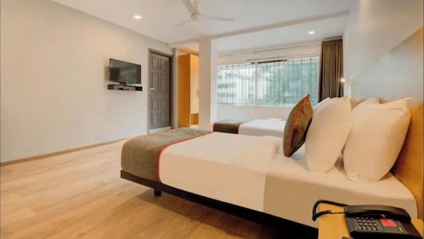 A hotel room with a bed, a bedside table with a telephone, a television mounted on the wall, and a window with curtains. - Maitrayee Hotel - Nagpur (Nagpur)