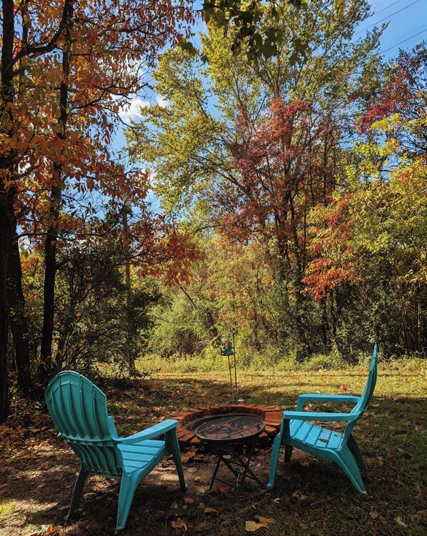 A circular fire pit with two turquoise chairs facing it, surrounded by trees with autumn foliage.