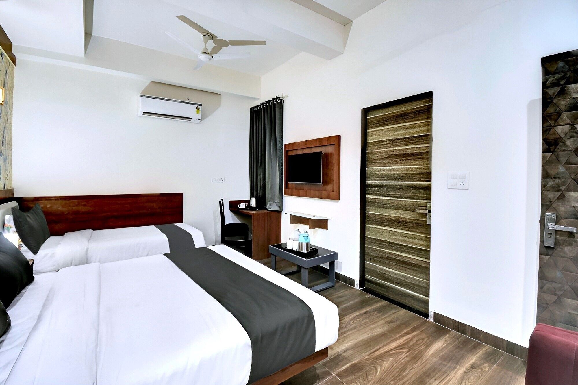 A hotel room with two beds, a ceiling fan, an air conditioning unit, a TV mounted on the wall, and a wooden door.