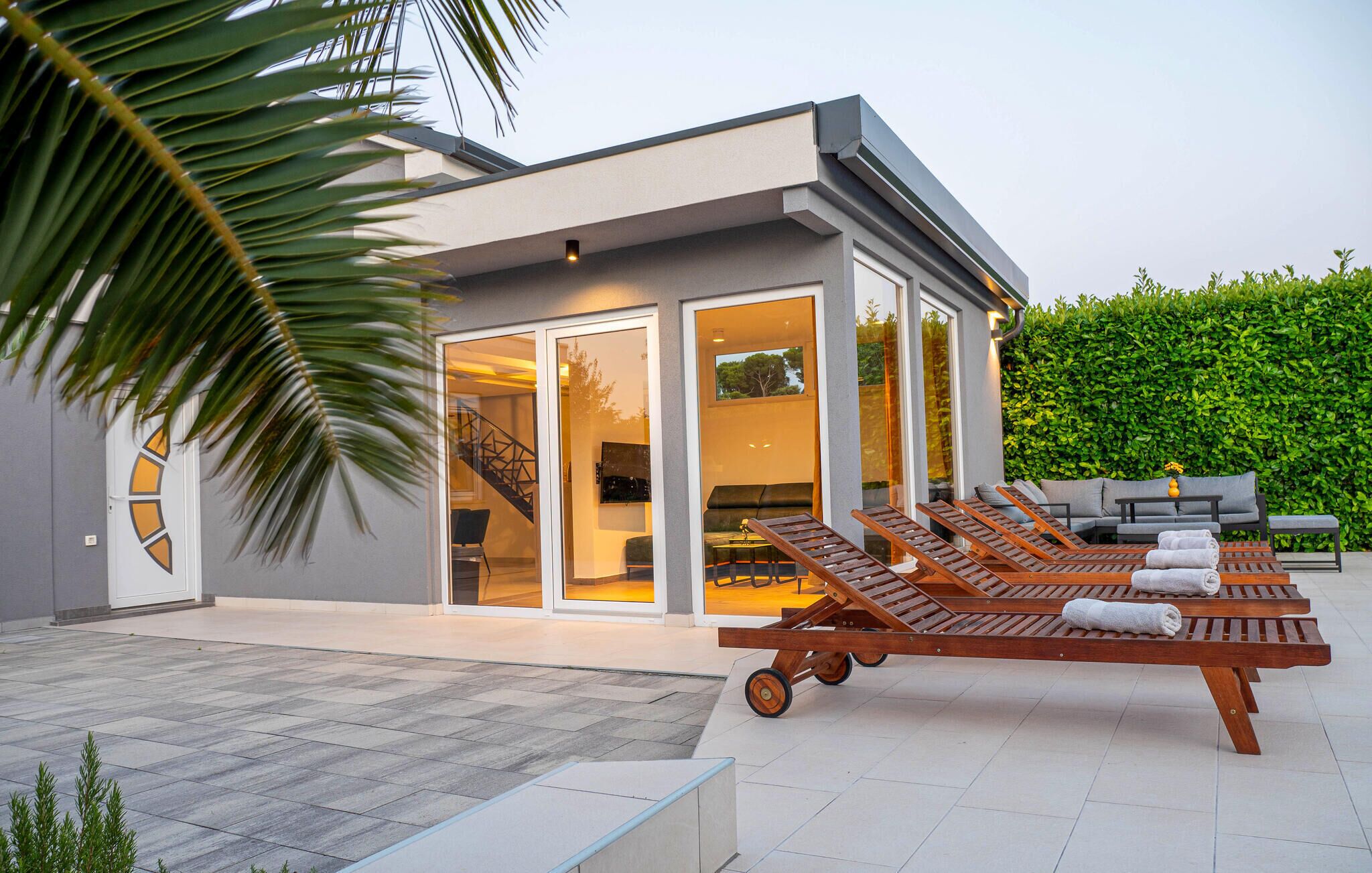 A modern house with a glass door leading to a living area, a patio with lounge chairs, and a neatly trimmed hedge.