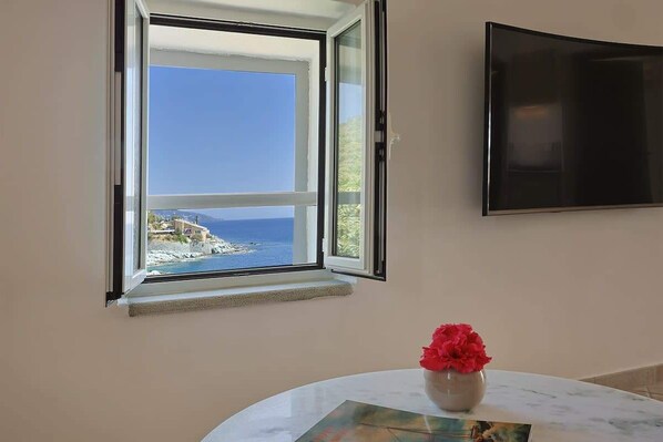 Habitación con una ventana grande que ofrece vistas al mar y a edificios, un televisor de pantalla plana en la pared y una mesita con un florero.
