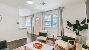 Een moderne woonkamer met een witte bank, twee fauteuils, een salontafel met een afstandsbediening, een grote plant en een roze deur op de achtergrond.