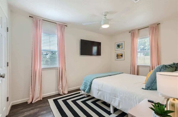 Ein Schlafzimmer mit einem Bett, Deckenventilator, Fernseher und einem Fenster mit Vorhängen.