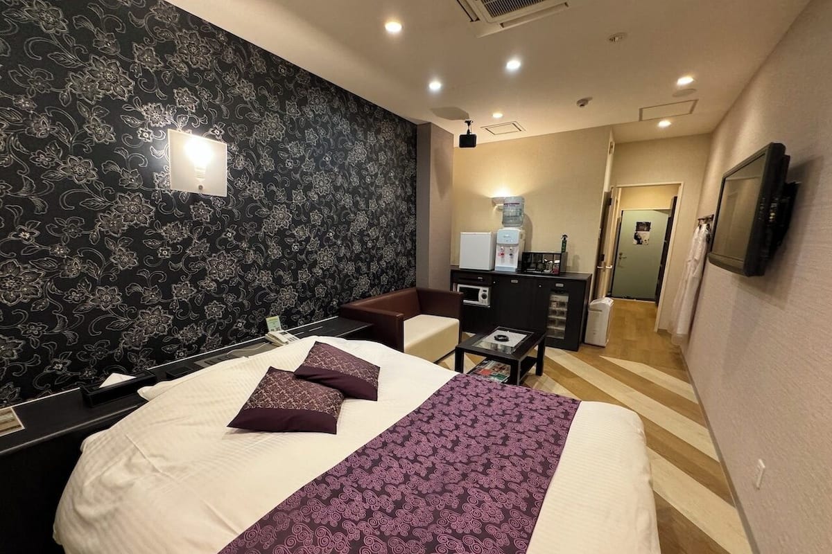 A hotel room with a large bed, a desk, a chair, a coffee machine, and a TV mounted on the wall.