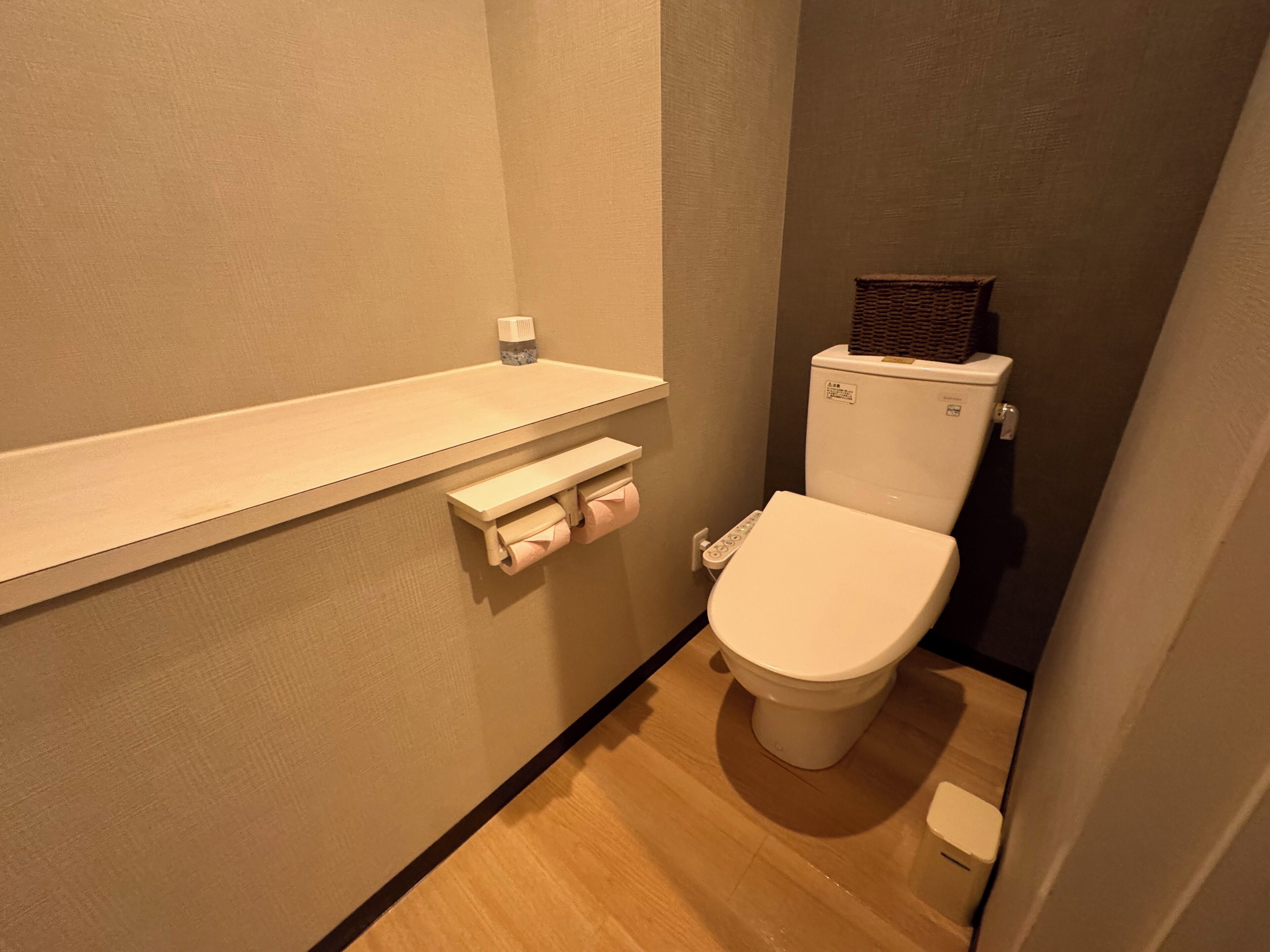 A small, compact bathroom with a toilet, a shelf with a bottle, and a toilet paper holder.