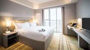 Quarto de hotel moderno com uma cama grande, mesa de cabeceira com abajur, escrivaninha com um laptop e televisão.