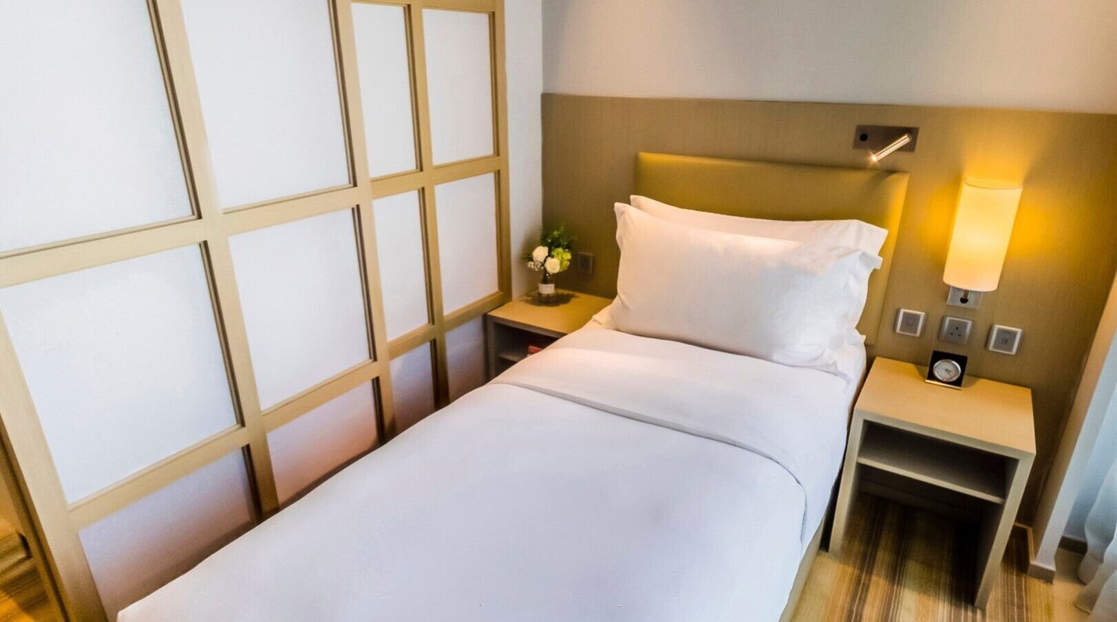 A hotel room with a bed, bedside tables, a nightstand with a lamp, and a wall-mounted light fixture.