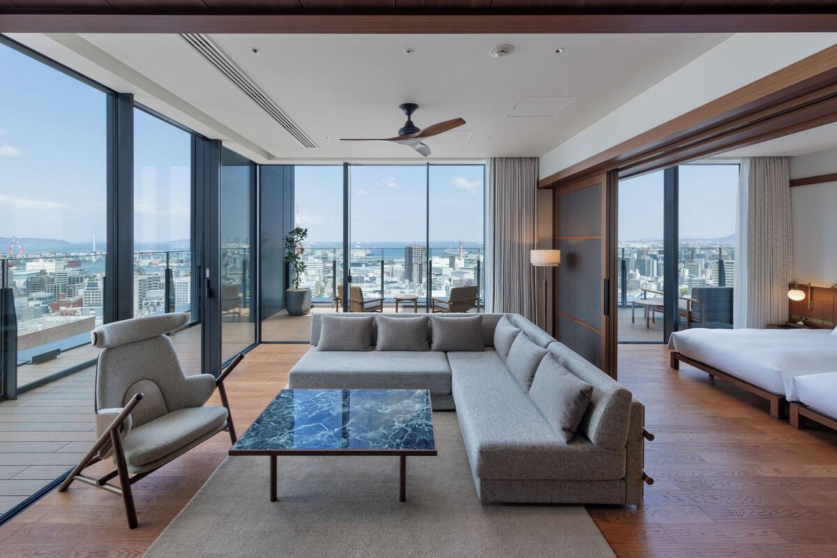 Une chambre d’hôtel moderne dotée d’un grand canapé d’angle, d’une table basse en marbre et offrant une vue sur la ville grâce à de grandes fenêtres du sol au plafond.