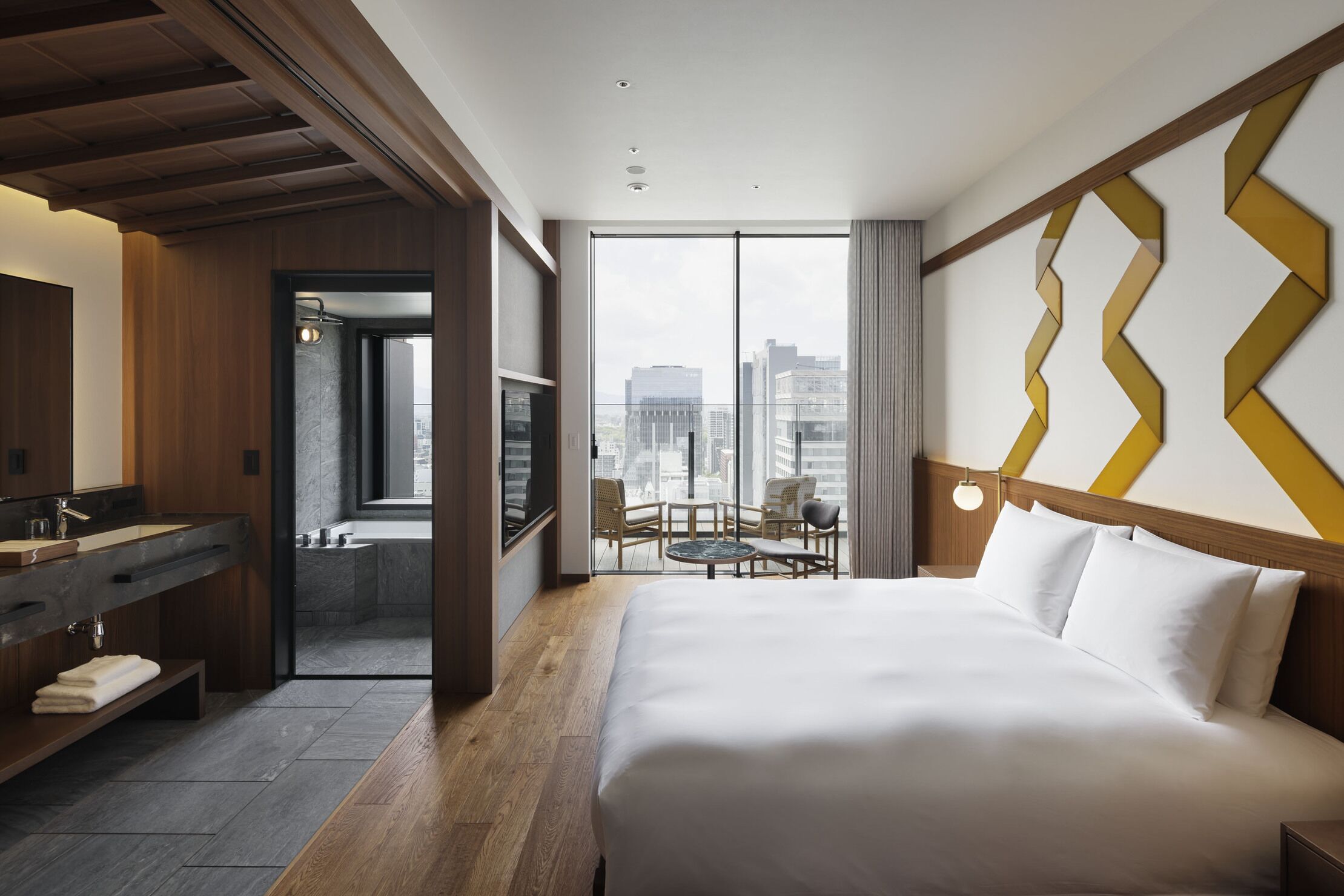 A hotel room with a large bed, a bathroom with a bathtub, a wooden vanity, and a city view.
