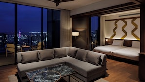 Une chambre d’hôtel moderne avec un canapé d’angle, une table basse en marbre, un lit avec une tête de lit et une vue sur la ville à travers de grandes fenêtres.