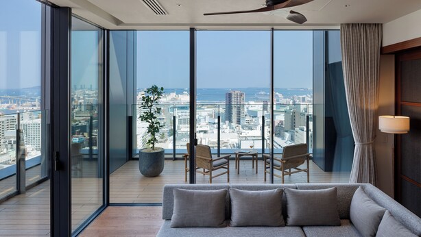 Une chambre d’hôtel moderne dotée d’un grand balcon, d’un canapé, de deux fauteuils, d’une plante en pot et offrant une vue sur le paysage urbain.