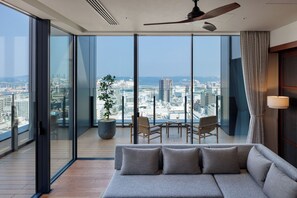 Une chambre d’hôtel moderne dotée d’un grand balcon, d’un canapé, de deux fauteuils, d’une plante en pot et offrant une vue sur le paysage urbain.