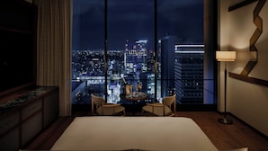 Une chambre d’hôtel dotée d’une grande fenêtre donnant sur la ville de nuit, avec un lit, deux chaises, une petite table sur laquelle se trouvent une bouteille et un verre, et un lampadaire.