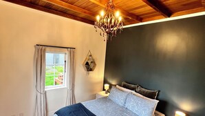 Ein Schlafzimmer mit Holzbalkendecke, einem Chandelier, einem Bett mit blau-weißer Bettdecke, einem Nachttisch mit Lampe und einem Fenster mit Vorhängen.