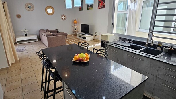 Et moderne køkken med en central ø, et spiseområde med stole, en sofa og et fjernsyn.