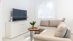 Un salon moderne avec un téléviseur à écran plat posé sur un meuble blanc, un canapé beige, une petite table ronde agrémentée d’une plante et une fenêtre ornée de rideaux légers.