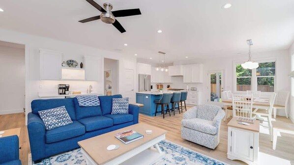 A modern living room with a blue sofa, a coffee table, and a white armchair. There is a kitchen area with white cabinets and a bar counter.