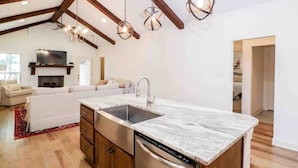 Una cocina moderna con encimera de mármol, electrodomésticos de acero inoxidable y horno empotrado. Hay un rincón comedor con sofá blanco y mesa de madera.