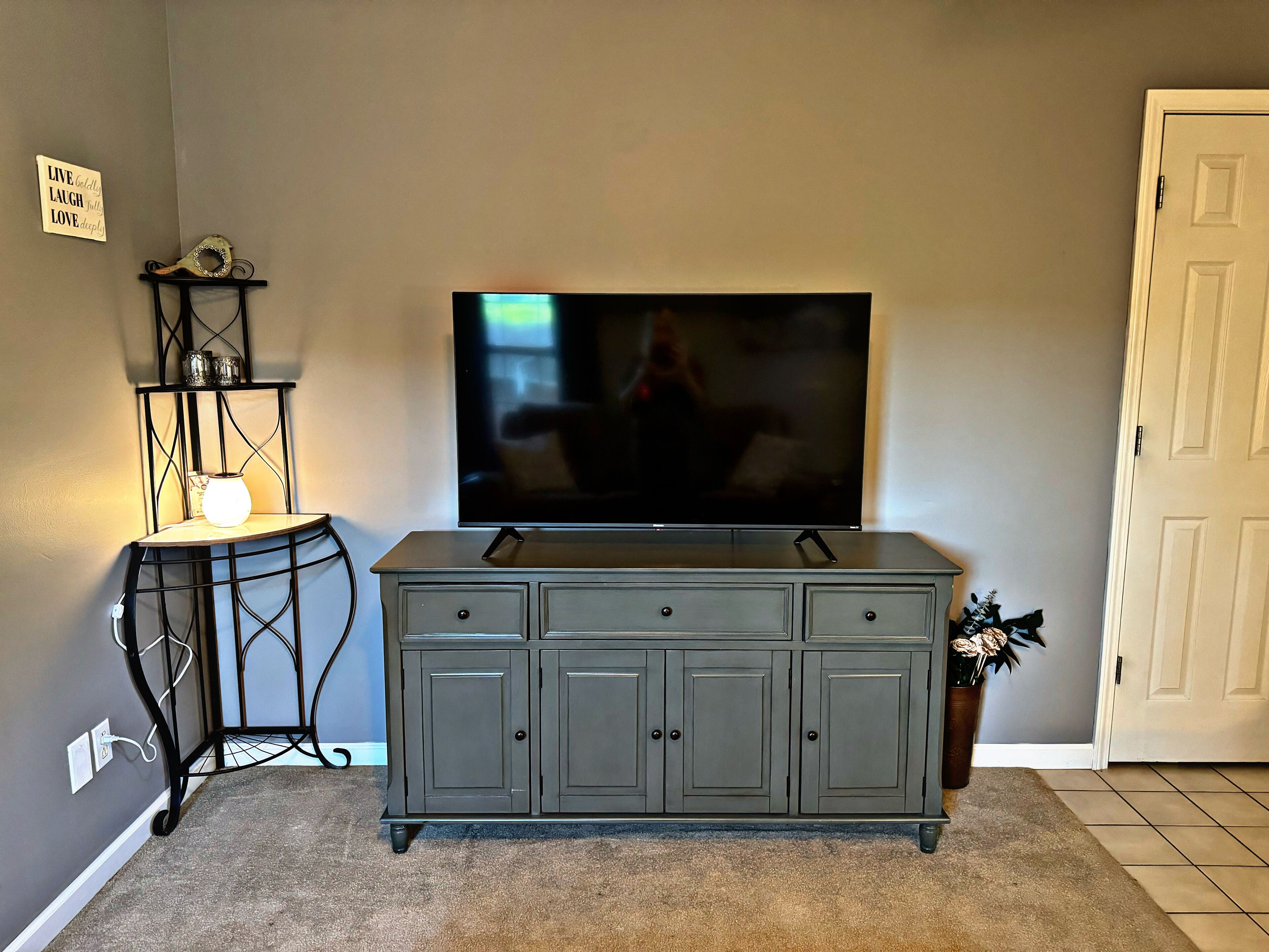A living room with a flat-screen TV mounted on the wall, a dark grey cabinet below, and a small table with a lamp and decorative items.
