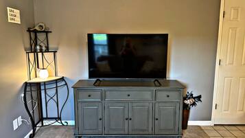A living room with a flat-screen TV mounted on the wall, a dark grey cabinet below, and a small table with a lamp and decorative items.