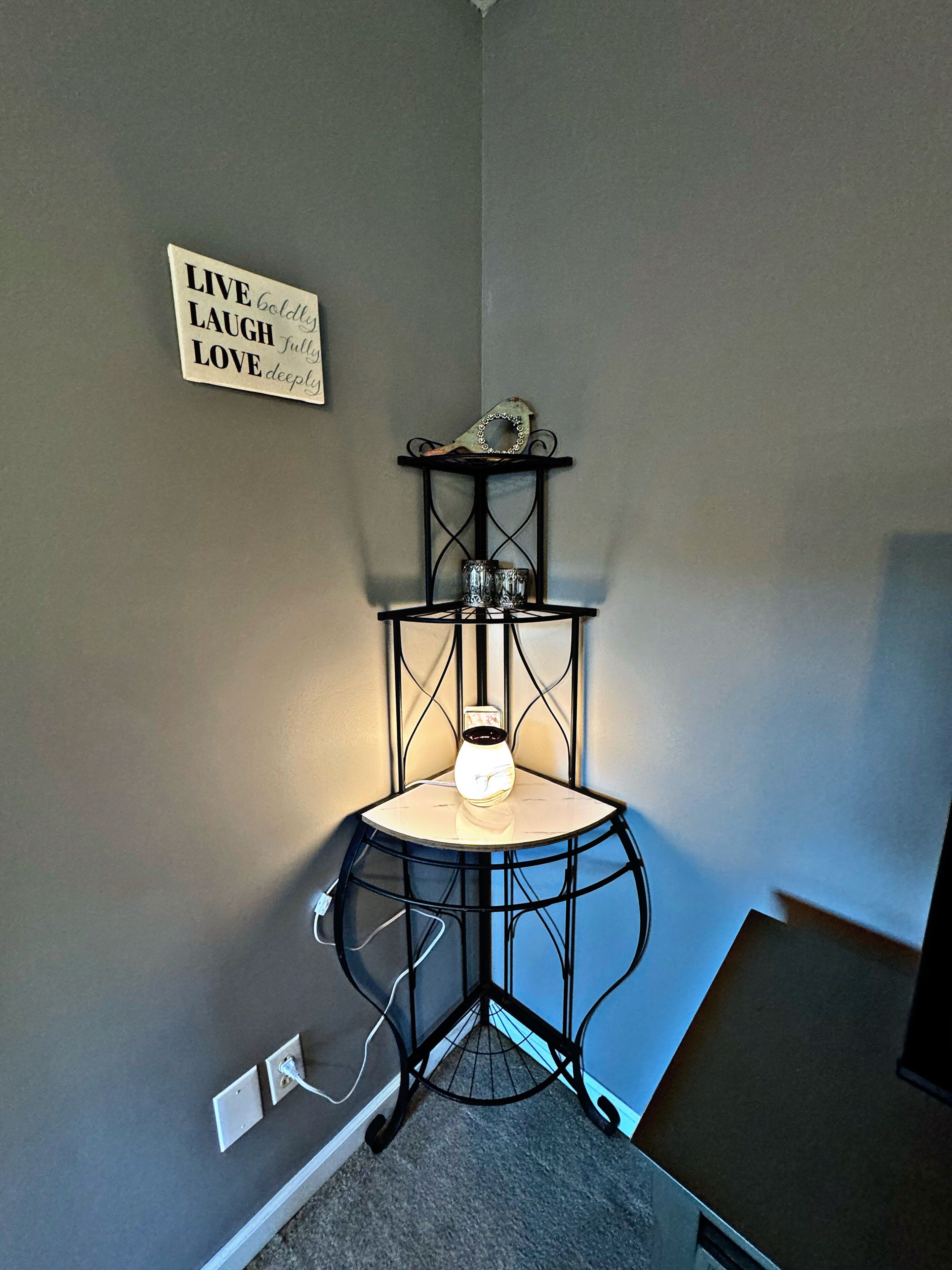 A corner room with a black metal shelf holding a vase and a decorative item, a wall-mounted sign with motivational text, and a power outlet.