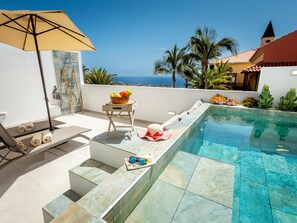 Zona de piscina con vistas al mar, una silla de descanso, una mesa pequeña con un cuenco de fruta y una sombrilla.