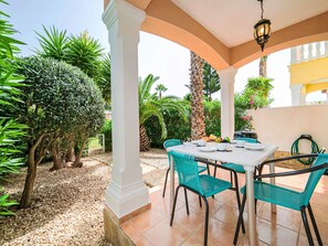 Eine Terrasse mit einem weißen Tisch und türkisblauen Stühlen, umgeben von Grünpflanzen und Palmen.