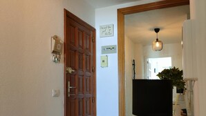 En gang med en trædør, et panelstyret termostat, en lyskontakt og et spejl der viser et rum med en plante, en lampe og et lille bord.