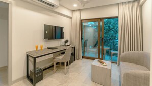 A modern living room with a flat-screen TV, a desk with a chair, a sofa, and a balcony with a view of trees. - Comfort Inn Elite Goa (Panaji)