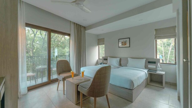 A hotel room with a large bed, a desk, two chairs, a ceiling fan, and a balcony with a table and chairs. - Comfort Inn Elite Goa (Panaji)