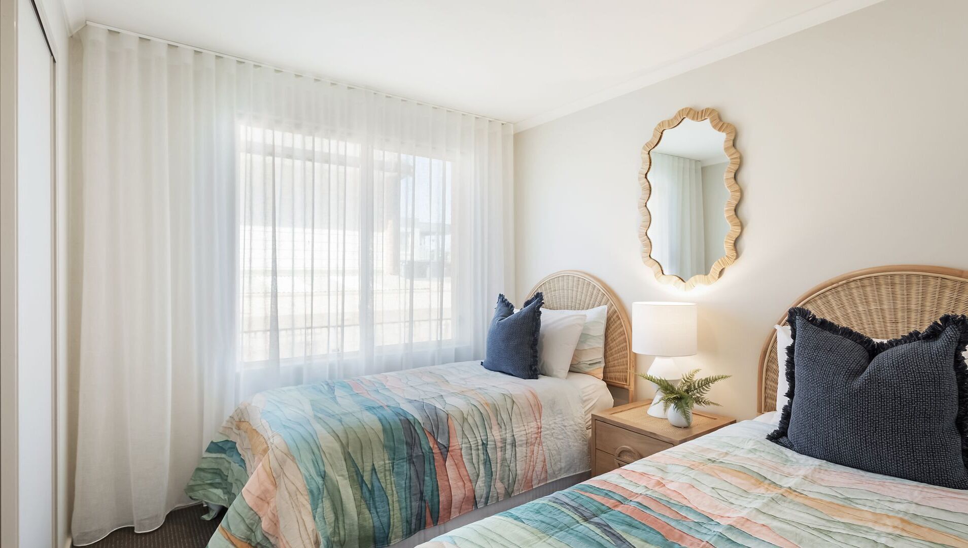 Habitación de hotel con dos camas, un ventanal con cortinas translúcidas, un espejo redondo y una mesita de noche con una lámpara y una planta.