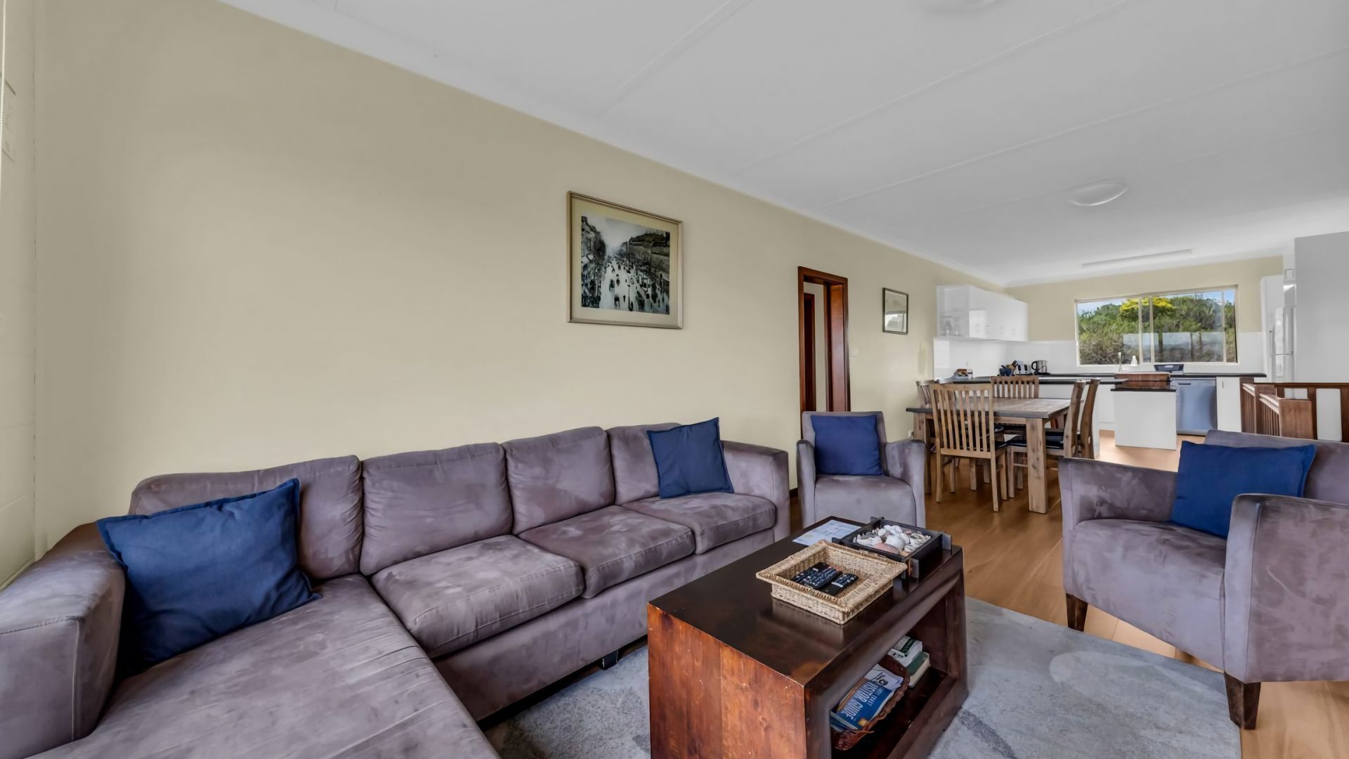 A modern living room with a grey sectional sofa, a wooden coffee table, and a dining area with wooden chairs and a white kitchen in the background.