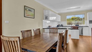 A modern kitchen with white cabinets, a wooden dining table, and a large window offering a view of greenery. - Cliffhanger - Pet Friendly! (Merimbula)