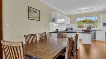 A modern kitchen with white cabinets, a wooden dining table, and a large window offering a view of greenery.